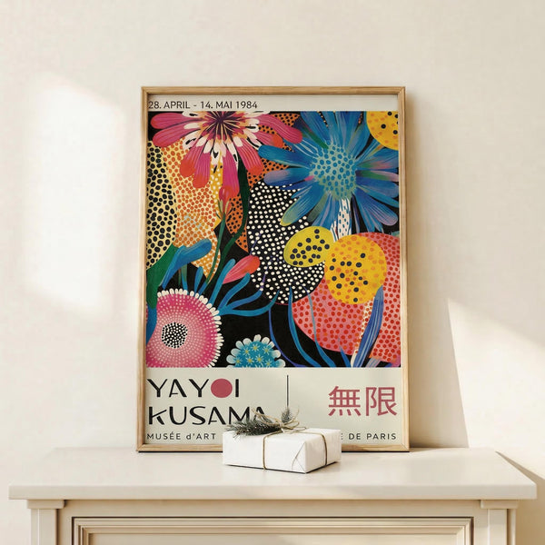 May include: Framed exhibition poster featuring vibrant floral artwork with colorful flowers in pink, blue, yellow, and orange against black background with polka dot patterns. Text shows "YAYOI KUSAMA" and "MUSEE d'ART DE PARIS" with dates "28. APRIL - 14. MAI 1984". Light wooden frame sits on white wooden surface next to small wrapped gift box with dried botanicals on top, against cream colored wall.