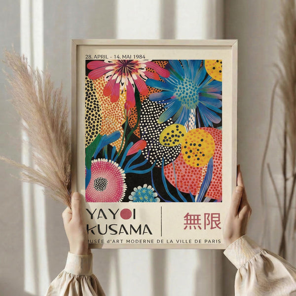 May include: Hands holding a framed colorful floral art print featuring vibrant pink, blue, and yellow abstract flowers with polka dot patterns against a black background. The poster shows exhibition text "28. APRIL - 14. MAI 1984" and "YAYOI KUSAMA" with "MUSÉE d'ART MODERNE DE LA VILLE DE PARIS" below. Light wooden frame, neutral beige wall background, dried pampas grass visible on the left side.