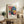 May include: Colorful Yayoi Kusama exhibition poster in light wood frame displayed on rustic wooden sideboard. The poster features vibrant floral designs with polka dots in pink, blue, yellow and orange against black background, with exhibition text below. Scene shows cozy living room with brown leather sofa, fern plant, ceramic vase, and stacked books creating warm, artistic home decor atmosphere.