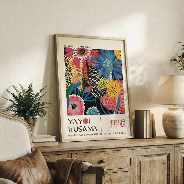 May include: Colorful Yayoi Kusama exhibition poster in light wood frame displayed on rustic wooden sideboard. The poster features vibrant floral designs with polka dots in pink, blue, yellow and orange against black background, with exhibition text below. Scene shows cozy living room with brown leather sofa, fern plant, ceramic vase, and stacked books creating warm, artistic home decor atmosphere.