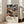 May include: A framed art poster leaning against a white wall next to a beige sofa in a bright room with large windows. The poster features swirling, colorful abstract patterns in blues, oranges, purples, and yellows with polka dots and flowing organic shapes. Text below reads "YAYOI KUSAMA" with Japanese characters and "永遠に続く愛 MUSÉE d'ART MODERNE DE LA VILLE DE PARIS" in a clean white border.