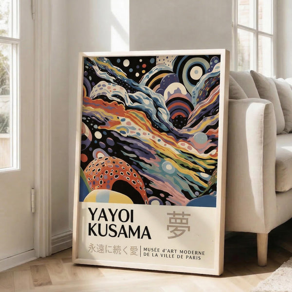 May include: A framed art poster leaning against a white wall next to a beige sofa in a bright room with large windows. The poster features swirling, colorful abstract patterns in blues, oranges, purples, and yellows with polka dots and flowing organic shapes. Text below reads "YAYOI KUSAMA" with Japanese characters and "永遠に続く愛 MUSÉE d'ART MODERNE DE LA VILLE DE PARIS" in a clean white border.