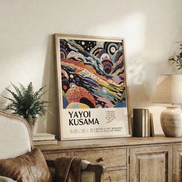 May include: Framed abstract art poster with swirling colorful patterns in purple, orange, and blue tones featuring polka dots and flowing organic shapes. Text reads "YAYOI KUSAMA" with Japanese characters and "MUSÉE d'ART MODERNE DE LA VILLE DE PARIS". Displayed on rustic wooden console table with books, ceramic vase, fern plant, and leather chair in warm neutral living room setting.