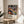 May include: Framed art poster featuring abstract swirling patterns in vibrant colors including orange, blue, purple, and yellow with polka dots and flowing organic shapes. Text reads "Yayoi Kusama" with Japanese characters and "Musée d'Art Moderne de la Ville de Paris". Natural wood frame sits on light wooden round side table next to beige upholstered chair with plant leaves visible, bright natural window light creating soft shadows on neutral wall.