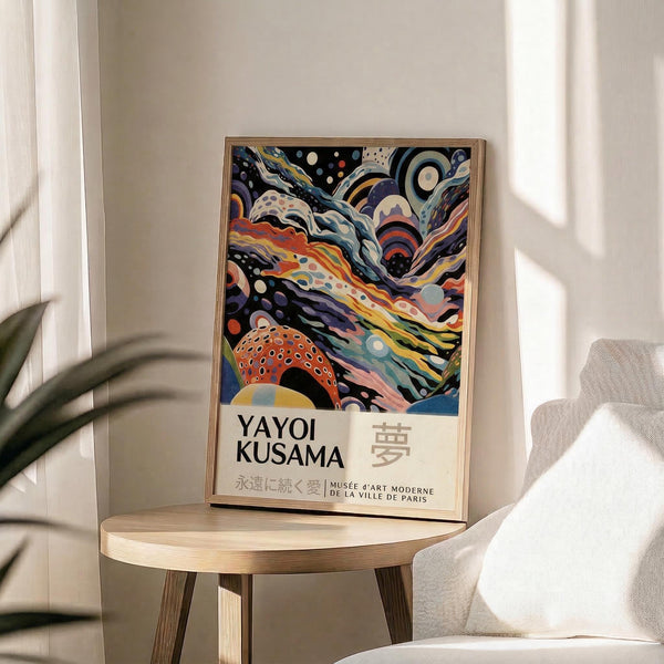May include: Framed art poster featuring abstract swirling patterns in vibrant colors including orange, blue, purple, and yellow with polka dots and flowing organic shapes. Text reads "Yayoi Kusama" with Japanese characters and "Musée d'Art Moderne de la Ville de Paris". Natural wood frame sits on light wooden round side table next to beige upholstered chair with plant leaves visible, bright natural window light creating soft shadows on neutral wall.