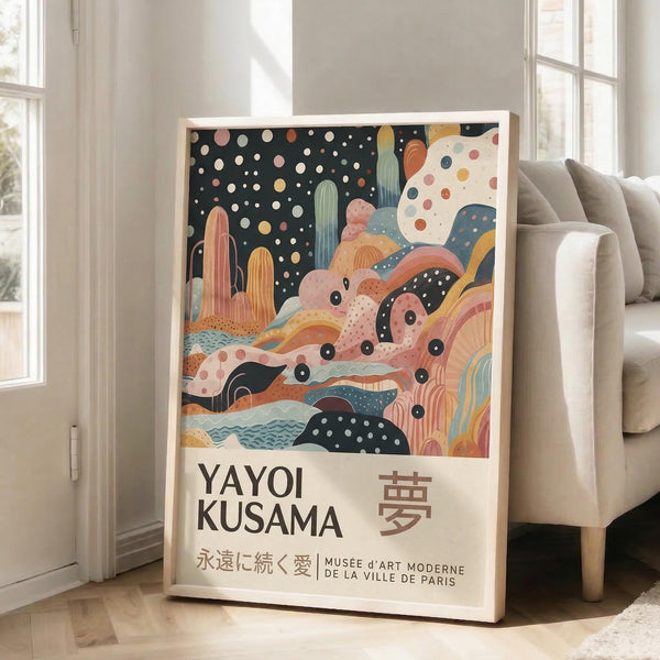 May include: Framed art print leaning against white wall next to beige sofa in bright living room. Artwork features whimsical desert landscape with colorful cacti, flowing abstract shapes in coral, teal, and yellow tones, polka dot patterns, and starry night sky. Text reads "YAYOI KUSAMA" with Japanese characters and "永遠に続く愛 MUSEE d'ART MODERNE DE LA VILLE DE PARIS" below the illustration.