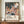 May include: Framed art poster displaying abstract colorful landscape with polka dots, cacti, and flowing organic shapes in pink, orange, blue, and cream tones against dark starry sky. Features "YAYOI KUSAMA" text and Japanese characters below artwork. Shown leaning against white wall on wooden console table next to small white ceramic vase and stacked books in bright room setting.