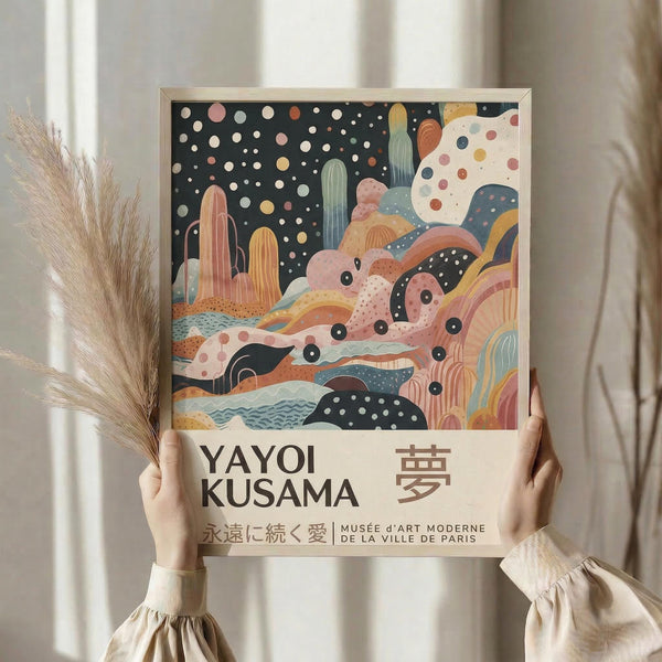 May include: Hands holding a framed art print featuring abstract colorful shapes, polka dots, and cactus forms in pink, orange, blue, and green tones against a dark starry background. Text reads "Yayoi Kusama" with Japanese characters. Dried pampas grass stems visible to the left against soft beige curtains in natural lighting.