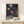 May include: Framed art print with navy blue background featuring colorful polka dot sunburst patterns in yellow, orange, and white. The poster shows text reading "YAYOI KUSAMA" and "MUSEE D'ART MODERNE DE LA VILLE DE PARIS" with Japanese characters. Displayed on white wooden surface beside small wrapped gift with evergreen sprig against cream wall.