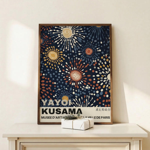 May include: Framed art print with navy blue background featuring colorful polka dot sunburst patterns in yellow, orange, and white. The poster shows text reading "YAYOI KUSAMA" and "MUSEE D'ART MODERNE DE LA VILLE DE PARIS" with Japanese characters. Displayed on white wooden surface beside small wrapped gift with evergreen sprig against cream wall.