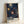 May include: Framed poster print leaning against white wall showing dark navy background with radiating sunburst patterns in cream, orange, and blue dots. Colorful firework-like designs spread across the composition. Text reads "YAYOI KUSAMA" and "MUSEE D'ART MODERNE DE LA VILLE DE PARIS" at bottom with Japanese characters. Light wooden frame surrounds the artwork in bright, minimally styled room setting.