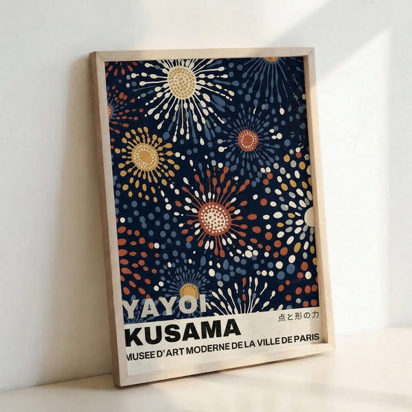 May include: Framed poster print leaning against white wall showing dark navy background with radiating sunburst patterns in cream, orange, and blue dots. Colorful firework-like designs spread across the composition. Text reads "YAYOI KUSAMA" and "MUSEE D'ART MODERNE DE LA VILLE DE PARIS" at bottom with Japanese characters. Light wooden frame surrounds the artwork in bright, minimally styled room setting.