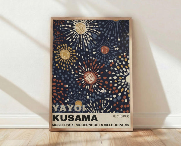 May include: Framed poster with dark blue background featuring abstract sunburst patterns in cream, orange, and gray dots radiating from circular centers. Text reads "Yayoi Kusama" and "Musée d'Art Moderne de la Ville de Paris" with Japanese characters. Wooden frame leans against white wall on light wood floor with soft shadows.