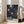 May include: Framed poster with dark navy background featuring colorful radiating sunburst patterns in yellow, orange, and white dots. Bold white text reads "YAYOI KUSAMA" and "MUSEE D'ART MODERNE DE LA VILLE DE PARIS" with Japanese characters. Light wood frame leans against white wall next to beige upholstered chair in bright, sunlit room with tall windows.
