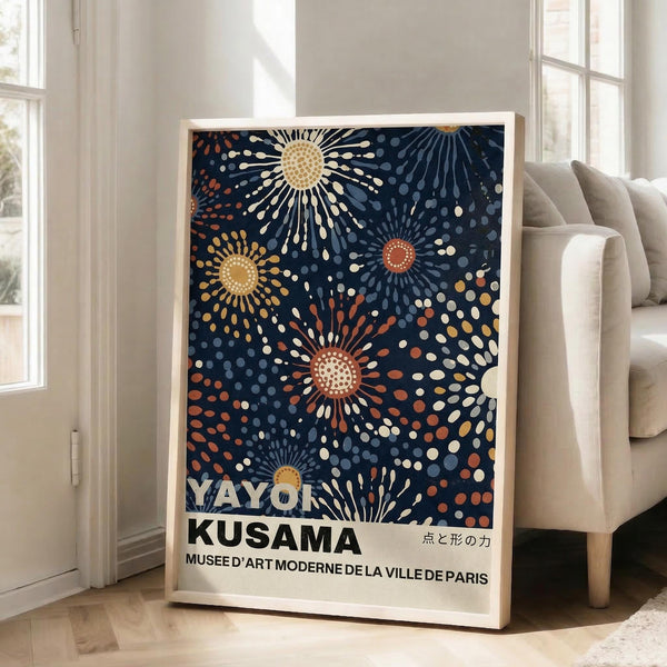 May include: Framed poster with dark navy background featuring colorful radiating sunburst patterns in yellow, orange, and white dots. Bold white text reads "YAYOI KUSAMA" and "MUSEE D'ART MODERNE DE LA VILLE DE PARIS" with Japanese characters. Light wood frame leans against white wall next to beige upholstered chair in bright, sunlit room with tall windows.
