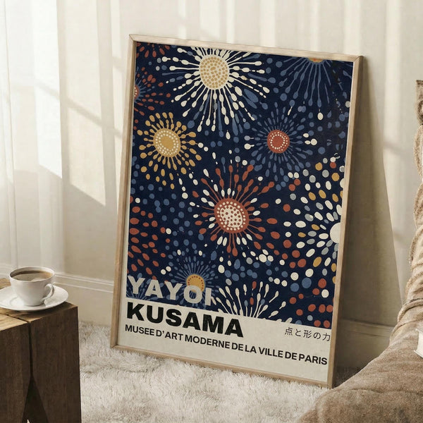 May include: Framed poster with navy blue background featuring colorful radiating dot patterns in cream, orange, and blue tones, displayed in light wood frame leaning against white wall next to coffee cup on wooden stool and cozy textured blanket in bright room with natural lighting