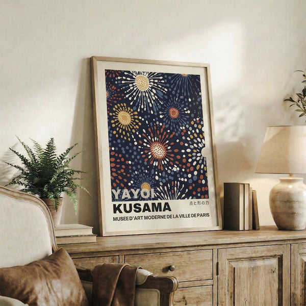 May include: Framed art print with dark blue background featuring colorful radiating sunburst patterns in yellow, orange, and white dots. Print displays "YAYOI KUSAMA" and museum text at bottom. Wooden frame sits on rustic wood sideboard next to fern plant, ceramic vase, and books in neutral-toned living room setting.