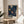 May include: Framed art print displaying abstract sunburst pattern in navy blue background with radiating dots in yellow, orange, white and coral colors, positioned on round wooden side table next to beige upholstered chair with decorative pillow, green plant leaves visible, soft natural lighting creating shadows on white wall