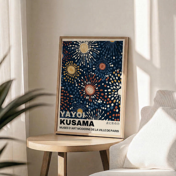May include: Framed art print displaying abstract sunburst pattern in navy blue background with radiating dots in yellow, orange, white and coral colors, positioned on round wooden side table next to beige upholstered chair with decorative pillow, green plant leaves visible, soft natural lighting creating shadows on white wall