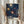 May include: Person holding framed art print featuring abstract sunburst pattern in navy blue background with radiating circles in yellow, orange, red, and white dots. Light wood frame, held against white curtained window with dried pampas grass visible to left side.