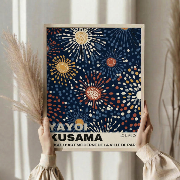 May include: Person holding framed art print featuring abstract sunburst pattern in navy blue background with radiating circles in yellow, orange, red, and white dots. Light wood frame, held against white curtained window with dried pampas grass visible to left side.