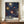 May include: Framed poster with navy blue background featuring colorful sunburst patterns in yellow, orange, and white dots radiating from central circles. Text reads "YAYOI KUSAMA" and "MUSEE D'ART MODERNE DE LA VILLE DE PARIS" with Japanese characters. Displayed on wooden sideboard next to white ceramic vase and books in elegant room with cream walls and white trim.