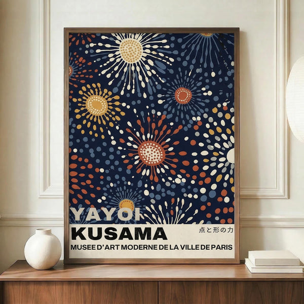 May include: Framed poster with navy blue background featuring colorful sunburst patterns in yellow, orange, and white dots radiating from central circles. Text reads "YAYOI KUSAMA" and "MUSEE D'ART MODERNE DE LA VILLE DE PARIS" with Japanese characters. Displayed on wooden sideboard next to white ceramic vase and books in elegant room with cream walls and white trim.