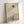 May include: Framed minimalist artwork leaning against white wall, featuring a bare black tree silhouette against layered beige and tan mountain shapes with a rust-colored circular sun above. The piece has a zen-like aesthetic with muted earth tones and is displayed in a thin natural wood frame on what appears to be a light wooden surface.