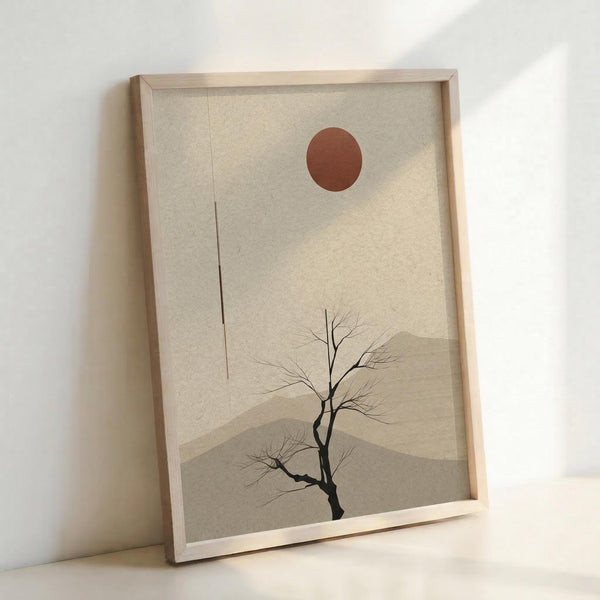 May include: Framed minimalist artwork leaning against white wall, featuring a bare black tree silhouette against layered beige and tan mountain shapes with a rust-colored circular sun above. The piece has a zen-like aesthetic with muted earth tones and is displayed in a thin natural wood frame on what appears to be a light wooden surface.