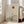 May include: Framed minimalist poster leaning against white wall beside beige armchair in bright living room. Artwork features bare black tree silhouette on textured beige background with rust-colored sun and layered mountain shapes. Natural wood frame, soft window lighting, hardwood floors visible.