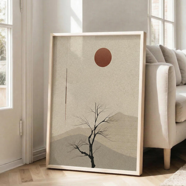 May include: Framed minimalist poster leaning against white wall beside beige armchair in bright living room. Artwork features bare black tree silhouette on textured beige background with rust-colored sun and layered mountain shapes. Natural wood frame, soft window lighting, hardwood floors visible.