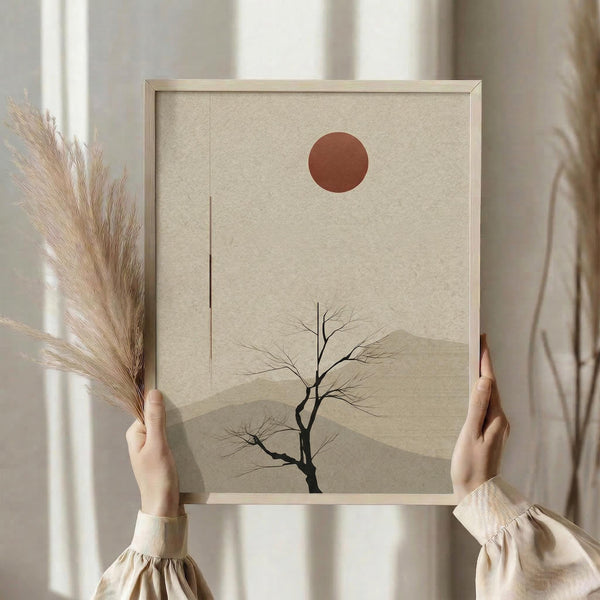 May include: Person in beige clothing holding framed minimalist artwork featuring a bare black tree silhouette against layered tan mountains with rust-colored sun above, displayed in light wood frame next to dried pampas grass in soft natural lighting near window with neutral curtains