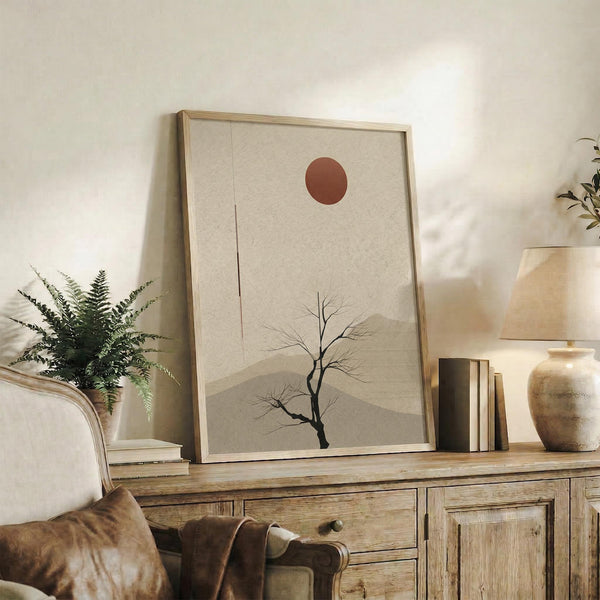 May include: Framed minimalist artwork featuring a bare tree silhouette against beige layered mountains with a rust-colored sun, displayed on a rustic wooden sideboard alongside fern plant, books, ceramic vase, and brown leather chair in warm neutral living room setting.