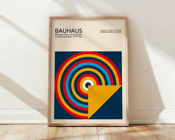 May include: Framed Bauhaus poster print in light wood frame, leaning against white wall on wooden floor. Features geometric design with colorful concentric circles in red, yellow, blue, and orange on dark blue background with yellow triangular shape. Modernist typography at top references the design school's history and locations.