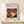 May include: Framed Bauhaus poster with geometric design featuring concentric circles in red, yellow, blue, and white with yellow triangular overlay on blue background, displayed on light wood surface next to small white gift box with greenery, against cream wall