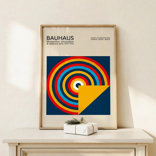 May include: Framed Bauhaus poster with geometric design featuring concentric circles in red, yellow, blue, and white with yellow triangular overlay on blue background, displayed on light wood surface next to small white gift box with greenery, against cream wall