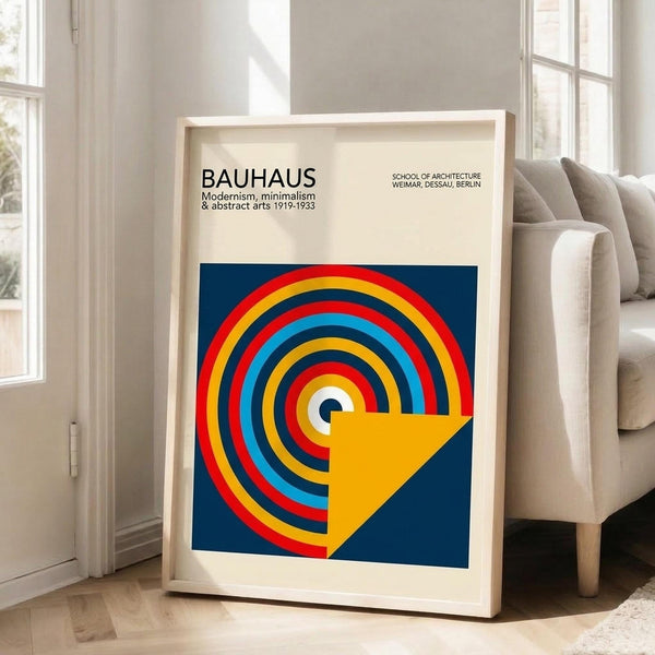 May include: Framed Bauhaus poster with geometric design featuring colorful concentric circles in red, yellow, blue, and white on navy background with yellow triangular shape. Light wood frame leaning against white wall next to beige sofa in bright living room with large windows and natural light casting shadows on wooden floor.