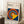 May include: Framed Bauhaus poster with colorful concentric circles in red, yellow, blue, and white on navy background, yellow geometric shape overlay. Light wood frame leaning against white wall corner next to wooden stool with white coffee cup and saucer, cream textured rug beneath.