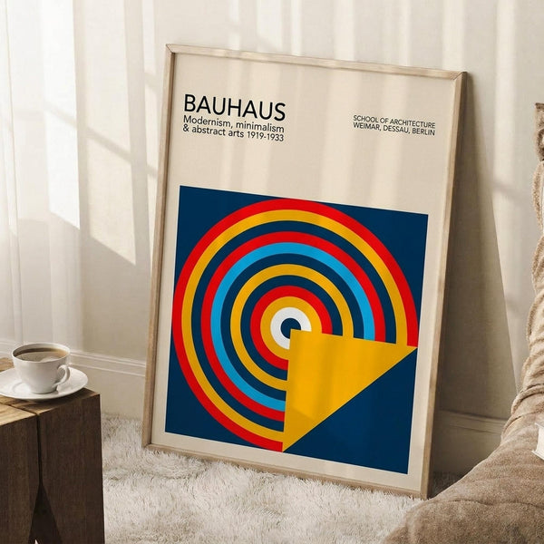 May include: Framed Bauhaus poster with colorful concentric circles in red, yellow, blue, and white on navy background, yellow geometric shape overlay. Light wood frame leaning against white wall corner next to wooden stool with white coffee cup and saucer, cream textured rug beneath.