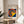 May include: Framed Bauhaus poster with colorful concentric circles in red, yellow, blue, and white on navy background, displayed on wooden round side table next to beige upholstered chair. Natural lighting creates soft shadows on white wall. Plant leaves visible in corner of minimalist modern room setting.