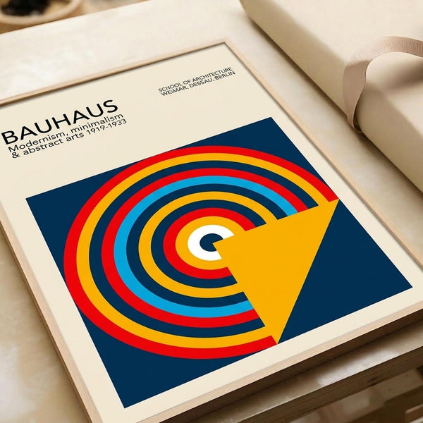 May include: Framed Bauhaus poster print featuring bold concentric circles in red, yellow, blue, and white on navy background with yellow triangular overlay. Modern minimalist design displayed in white matted frame on cream surface beside rolled papers, showcasing the geometric art movement's characteristic clean lines and primary color palette.