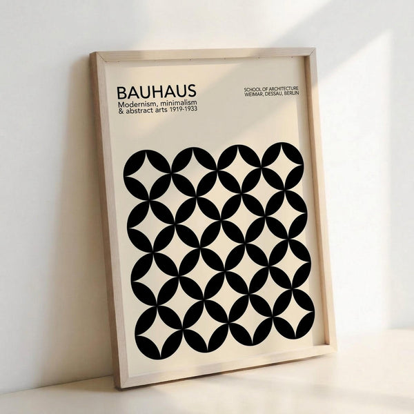May include: Framed poster with cream background featuring black geometric pattern of overlapping circles creating star-like shapes. Text reads "BAUHAUS Modernism, minimalism & abstract arts 1919-1933" and "SCHOOL OF ARCHITECTURE WEIMAR, DESSAU, BERLIN." Light wood frame leans against white wall, showing natural lighting and shadows.