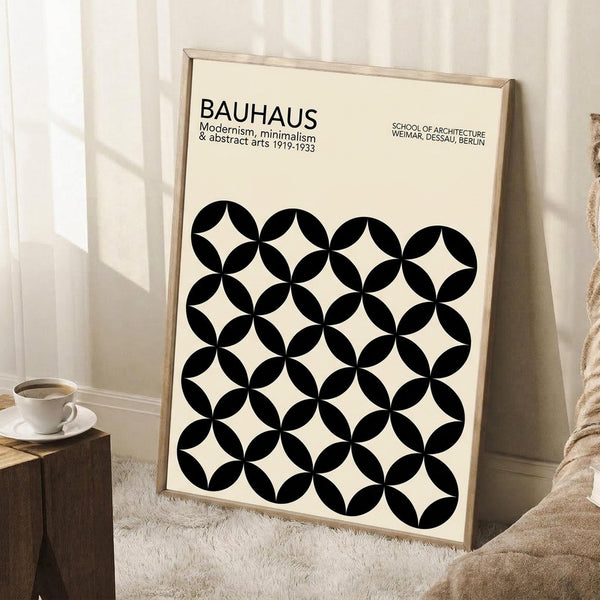 May include: A framed poster with cream background featuring black geometric circles in an overlapping pattern, creating four-pointed star shapes between them. Text reads "BAUHAUS Modernism, minimalism & abstract arts 1919-1933" and "SCHOOL OF ARCHITECTURE WEIMAR, DESSAU, BERLIN." The wooden-framed artwork leans against a white wall near a beige armchair, with a coffee cup on a wooden side table and soft natural lighting.