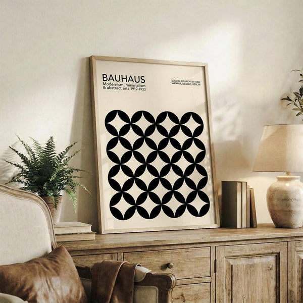 May include: Framed Bauhaus poster with geometric black circles pattern on cream background, displayed on rustic wooden sideboard alongside fern plant, ceramic vase, and brown leather chair in neutral-toned living room setting.