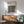 May include: Framed Bauhaus poster with black and white geometric circle pattern hanging above wooden sideboard. Mid-century modern living room setting with sage green upholstered chair, round wooden coffee table, and white ceramic table lamp. Natural sunlight creates shadows on white wall. Wood frame matches furniture tones.