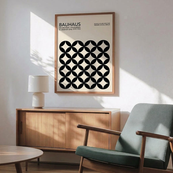 May include: Framed Bauhaus poster with black and white geometric circle pattern hanging above wooden sideboard. Mid-century modern living room setting with sage green upholstered chair, round wooden coffee table, and white ceramic table lamp. Natural sunlight creates shadows on white wall. Wood frame matches furniture tones.