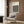 May include: Cream poster with black geometric circle pattern and "BAUHAUS" text hanging on wood-paneled wall above beige upholstered armchair with patterned pillow. Scene shows modern reading nook with brass floor lamp, dark round side table with open book, and white curtains. Warm neutral tones throughout minimalist interior setting.
