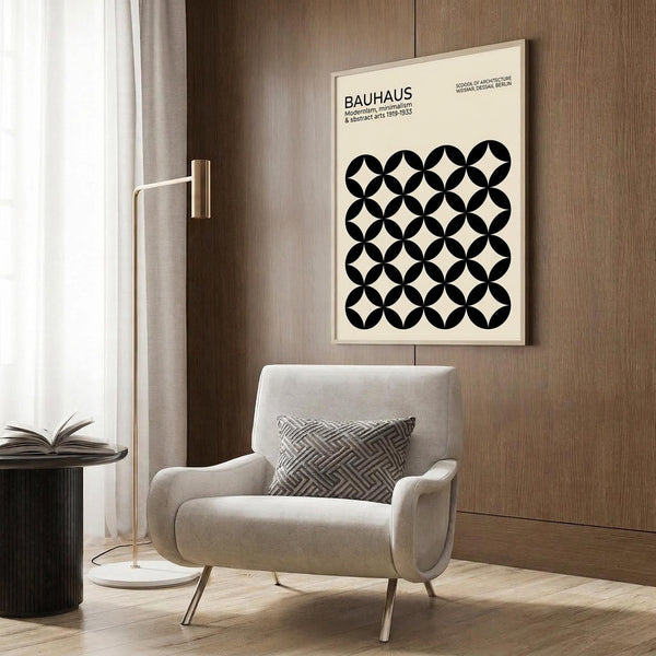 May include: Cream poster with black geometric circle pattern and "BAUHAUS" text hanging on wood-paneled wall above beige upholstered armchair with patterned pillow. Scene shows modern reading nook with brass floor lamp, dark round side table with open book, and white curtains. Warm neutral tones throughout minimalist interior setting.