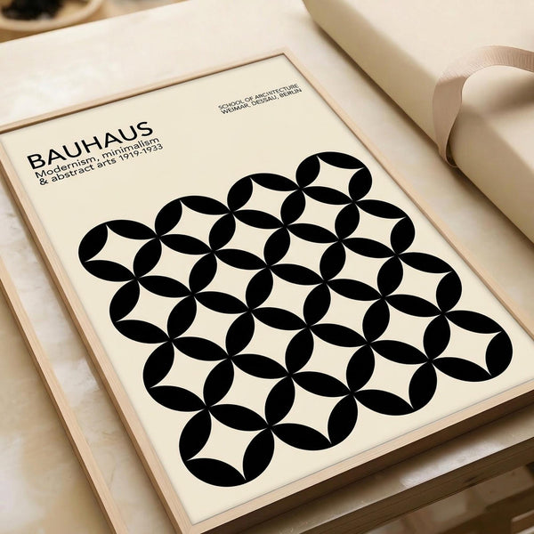 May include: Framed Bauhaus poster with black geometric circle pattern on cream background, displayed on light wood surface next to rolled paper tube. Modern minimalist design features overlapping circles creating repeating motif. Clean typography references the German design school's modernist aesthetic from 1919-1933.