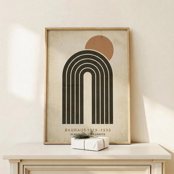 May include: Framed Bauhaus-inspired poster with geometric design featuring concentric arches in dark green and cream colors, topped by a terracotta circle resembling a sun. The minimalist artwork sits on a light wooden console against a cream wall, with a small white wrapped gift beside it. The print has vintage-style text at bottom and warm, natural lighting creates soft shadows.